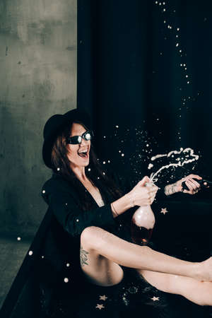 girl in a black jacket and hat posing in a black bath. A bottle of rose champagne wine in hand. Stylish sunglasses. Long naked sexy legs. splashes of champagneの写真素材