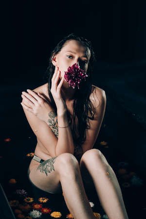 Young pretty brunette woman with tattoos in bath with water and flowers. Naked model during an unusual photo shoot in a black bath. Flower in the mouthの写真素材