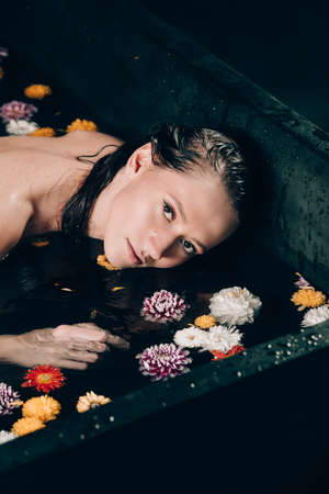 Young pretty brunette woman with tattoos in bath with water and flowers. Naked model during an unusual photo shoot in a black bath.の写真素材