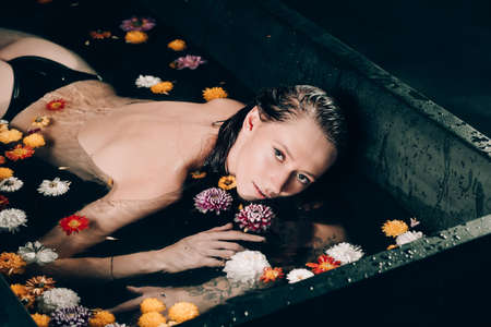 Young pretty brunette woman with tattoos in bath with water and flowers. Naked model during an unusual photo shoot in a black bath.の写真素材