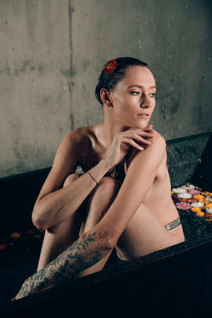 Young pretty brunette woman with tattoos in bath with water and flowers. Naked model during an unusual photo shoot in a black bath.の写真素材