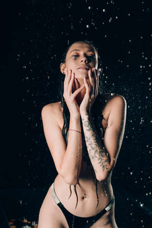 Young pretty brunette woman with tattoos in bath with water and flowers. Naked model during an unusual photo shoot in a black bath.の写真素材