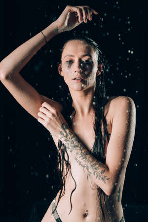 Young pretty brunette woman with tattoos in bath with water and flowers. Naked model during an unusual photo shoot in a black bath.の写真素材