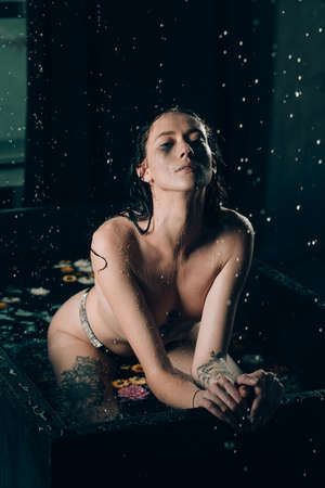 Young pretty brunette woman with tattoos in bath with water and flowers. Naked model during an unusual photo shoot in a black bath.の写真素材