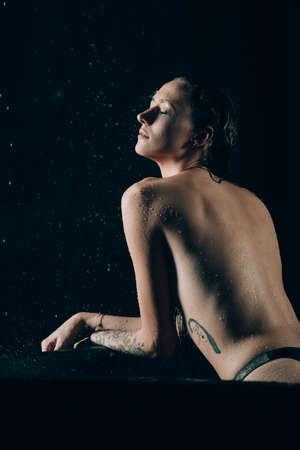 Young pretty brunette woman with tattoos in bath with water and flowers. Naked model during an unusual photo shoot in a black bath.の写真素材