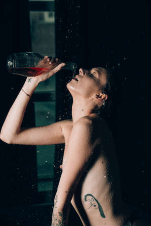Young pretty brunette woman with tattoos in bath with water and flowers. Naked model during an unusual photo shoot in a black bath drinking wineの写真素材