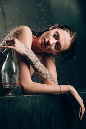 Young pretty brunette woman with tattoos in a bath. Naked model during an unusual photo shoot in a black bath drinking wineの写真素材