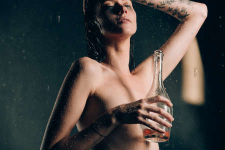Young pretty brunette woman with tattoos in bath with water and flowers. Naked model during an unusual photo shoot in a black bath drinking wineの写真素材
