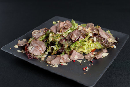 Salad with smoked duck meat, sweet and sour sauce with almonds flakes on a black ceramic plateの写真素材