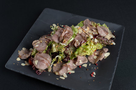 Salad with smoked duck meat, sweet and sour sauce with almonds flakes on a black ceramic plateの写真素材