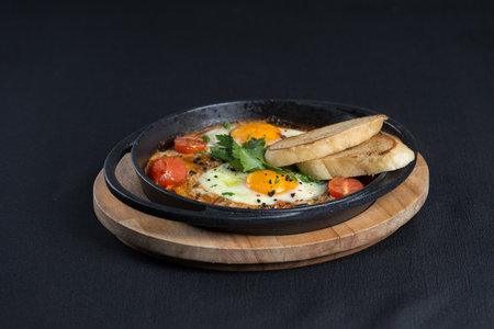 healthy breakfast shakshuka - fried eggs, onion, bell pepper, tomatoes and spices in cast iron stewpan on old wooden plank, view from hight angle. Black tableclothの写真素材