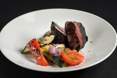 veal tenderloin in ashes with grilled vegetables on a white plate on a black tablecloth.の写真素材