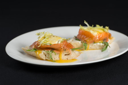 A delicious eggs benedict with lightly salted salmon and sauce on a white plate on a black tablecloth. Top viewの写真素材