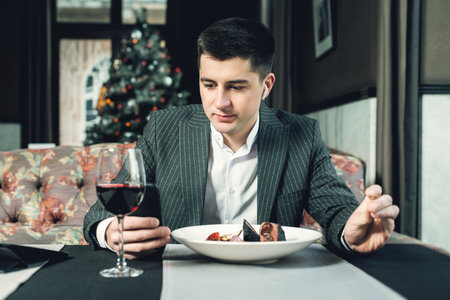 A young and attractive man uses his phone while eating veal tenderloin in ashes with grilled vegetables in an indoor restaurant. Business person work indoor conceptの写真素材