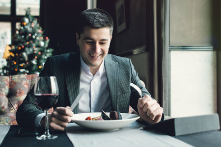 A young and attractive man uses his digital devises while eating veal tenderloin in ashes with grilled vegetables in an indoor restaurant. Business person work indoor conceptの写真素材