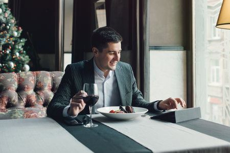 A young and attractive man uses his phone while eating veal tenderloin in ashes with grilled vegetables in an indoor restaurant. Business person work indoor conceptの写真素材