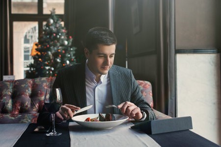 A young and attractive man uses his phone while eating veal tenderloin in ashes with grilled vegetables in an indoor restaurant. Business person work indoor conceptの写真素材