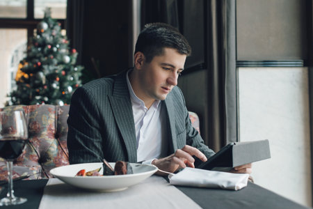 A young and attractive man uses his phone while eating veal tenderloin in ashes with grilled vegetables in an indoor restaurant. Business person work indoor conceptの写真素材