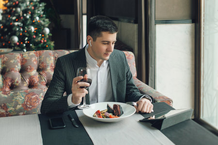 A young and attractive man uses his phone while eating veal tenderloin in ashes with grilled vegetables in an indoor restaurant. Business person work indoor conceptの写真素材