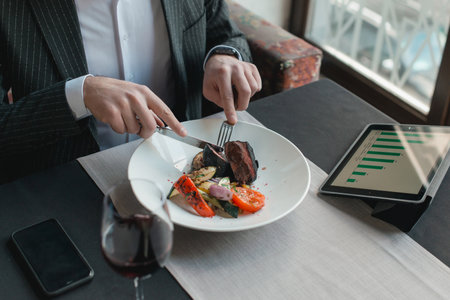 A young and attractive man uses his phone while eating veal tenderloin in ashes with grilled vegetables in an indoor restaurant. Business person work indoor conceptの写真素材