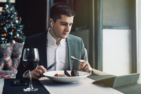 A young and attractive man uses his phone while eating veal tenderloin in ashes with grilled vegetables in an indoor restaurant. Business person work indoor conceptの写真素材