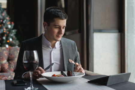 A young and attractive man uses his phone while eating veal tenderloin in ashes with grilled vegetables in an indoor restaurant. Business person work indoor conceptの写真素材