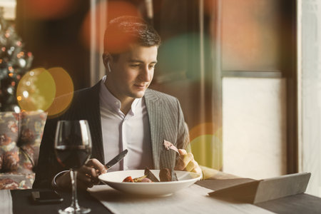 A young and attractive man uses his phone while eating veal tenderloin in ashes with grilled vegetables in an indoor restaurant. Business person work indoor conceptの写真素材