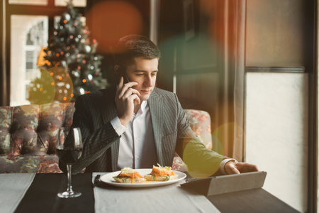 A young and attractive man uses his phone while eating veal tenderloin in ashes with grilled vegetables in an indoor restaurant. Business person work indoor conceptの写真素材