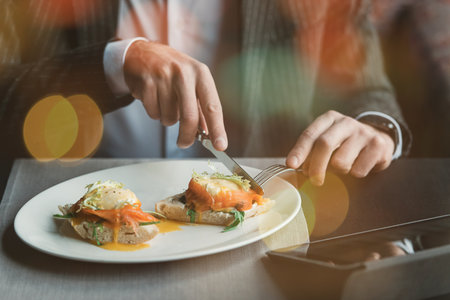 A young and attractive man uses his phone while eating delicious eggs benedict with lightly salted salmon and sauce in an indoor restaurant. Business person work indoor conceptの写真素材