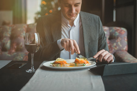 A young and attractive man uses his phone while eating veal tenderloin in ashes with grilled vegetables in an indoor restaurant. Business person work indoor conceptの写真素材