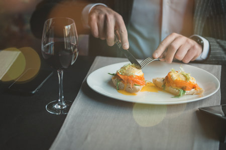 A young and attractive man uses his phone while eating delicious eggs benedict with lightly salted salmon and sauce in an indoor restaurant. Business person work indoor conceptの写真素材