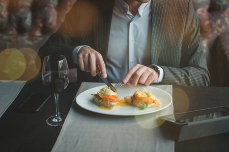 A young attractive man eating cottage cheese pancakes with sour cream and almond flakes and sea buckthorn-orange sauce in an indoor restaurant. Business person work indoor conceptの写真素材