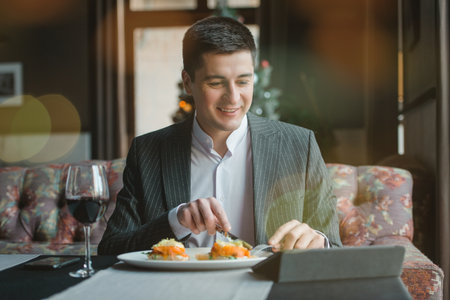 A young attractive man eating cottage cheese pancakes with sour cream and almond flakes and sea buckthorn-orange sauce in an indoor restaurant. Business person work indoor conceptの写真素材