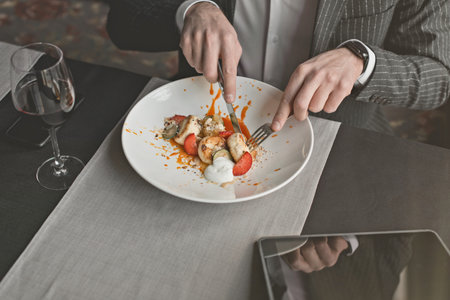 A young attractive man eating cottage cheese pancakes with sour cream and almond flakes and sea buckthorn-orange sauce in an indoor restaurant. Business person work indoor conceptの写真素材