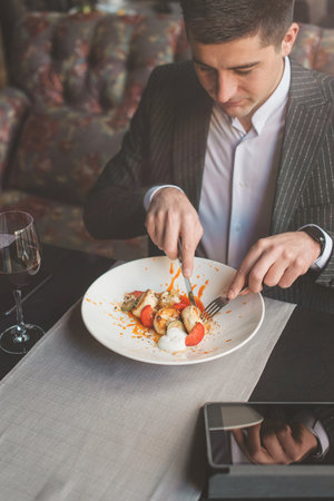 A young attractive man eating cottage cheese pancakes with sour cream and almond flakes and sea buckthorn-orange sauce in an indoor restaurant. Business person work indoor conceptの写真素材