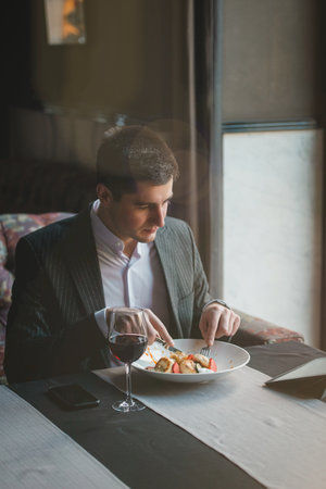 A young attractive man eating cottage cheese pancakes with sour cream and almond flakes and sea buckthorn-orange sauce in an indoor restaurant. Business person work indoor conceptの写真素材