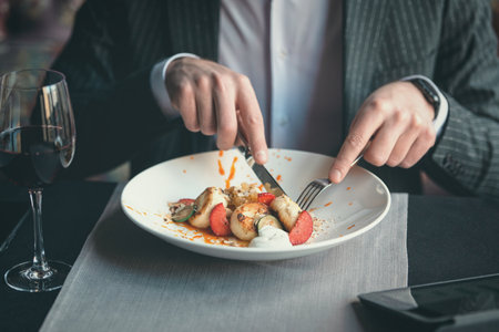 A young attractive man eating cottage cheese pancakes with sour cream and almond flakes and sea buckthorn-orange sauce in an indoor restaurant. Business person work indoor conceptの写真素材