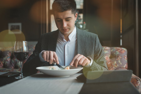 A young and attractive man uses his tablet while eating turkey fillet roll and pea puree in an indoor restaurant. Business person work indoor conceptの写真素材