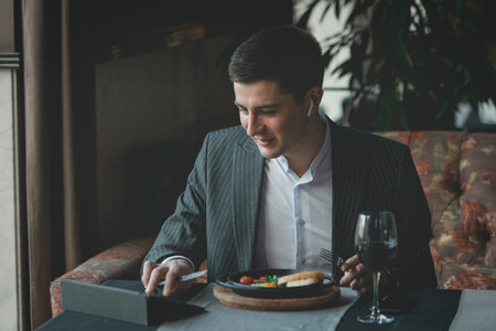 man uses his tablet while eating healthy breakfast shakshuka - fried eggs, tomatoes and spices in cast iron stewpan on old wooden plank in an indoor restaurant.の写真素材