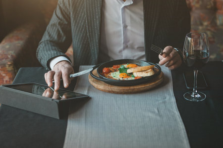 man uses his tablet while eating healthy breakfast shakshuka - fried eggs, tomatoes and spices in cast iron stewpan on old wooden plank in an indoor restaurant.の写真素材