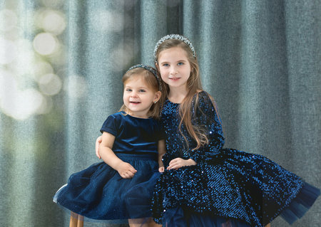 two cute baby girls sisters with blue eyes wear dark blue dresses sit on a decorative bench and hugの写真素材