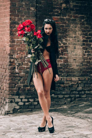Elegant beautiful brunette woman in red shorts and black coat posing near brick wall with a big bouquet of red roses. Birthday. Valentines day. Selective focus, filmgrainの写真素材