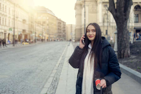 Stylish Caucasian woman in a coat stands and drinks coffee on the street. Urban style and fashionable girl with a red paper cup of coffee.の写真素材