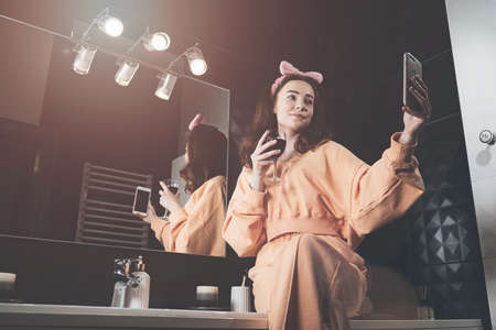 Attractive young woman in a bathroom communicating on a smartphone and drinking wine. Relaxation and leisure concept.の写真素材