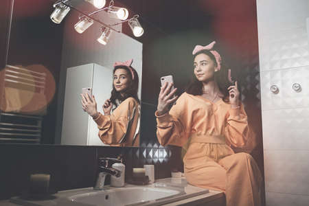 beautiful young woman communicating by the phone and doing her hair in a stylish bathroom. dressed in an orange sports suit and a bowの写真素材