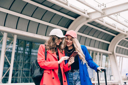 Two happy girls using smartphone checking flight or online check-in at airport together, with luggage. Air travel, summer holidayの写真素材