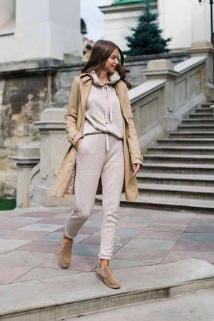 Portrait of fashionable women in beige sports suit, trench coat and stylish suede loafer posing on the stone stairsの写真素材