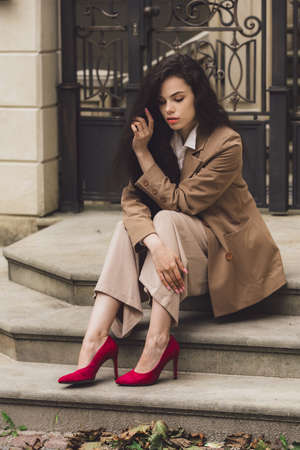 Close up portrait of young beautiful woman with long brunette curly hair posing against building background. sitting on the stairs. bright red high heeled shoesの写真素材