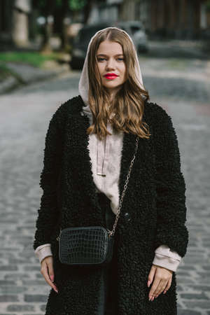 Close-up photo of young beautiful woman in black fur coat, jeanse and shoes posing on streetの写真素材