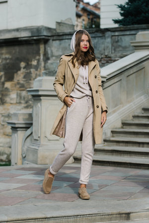 Portrait of fashionable women in beige sports suit, trench coat and stylish suede loafer posing on the stone stairsの写真素材